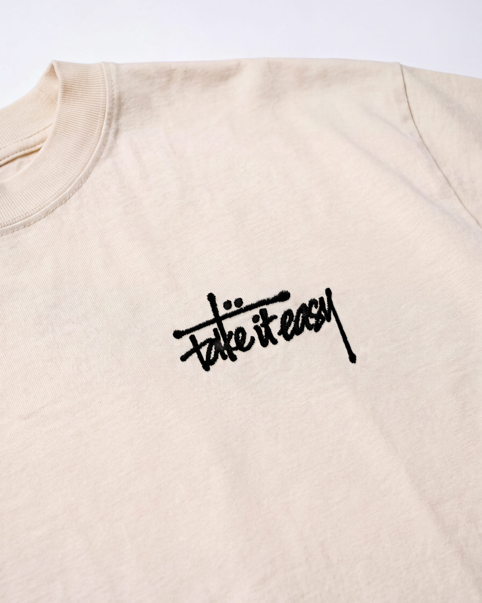Close-up of the embroidered “take it easy” mark on the chest of the cream tee